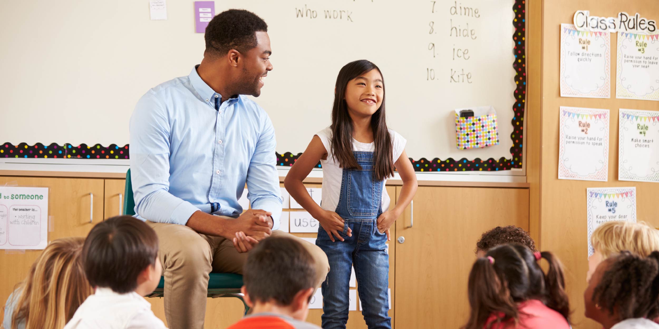 A student standing in front of the class with the teacher next to her